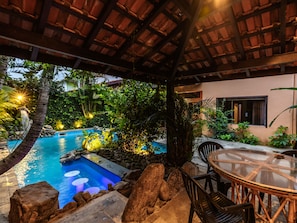 Zona de piscina con mesa y sillas de vidrio, rodeada de rocas y plantas.