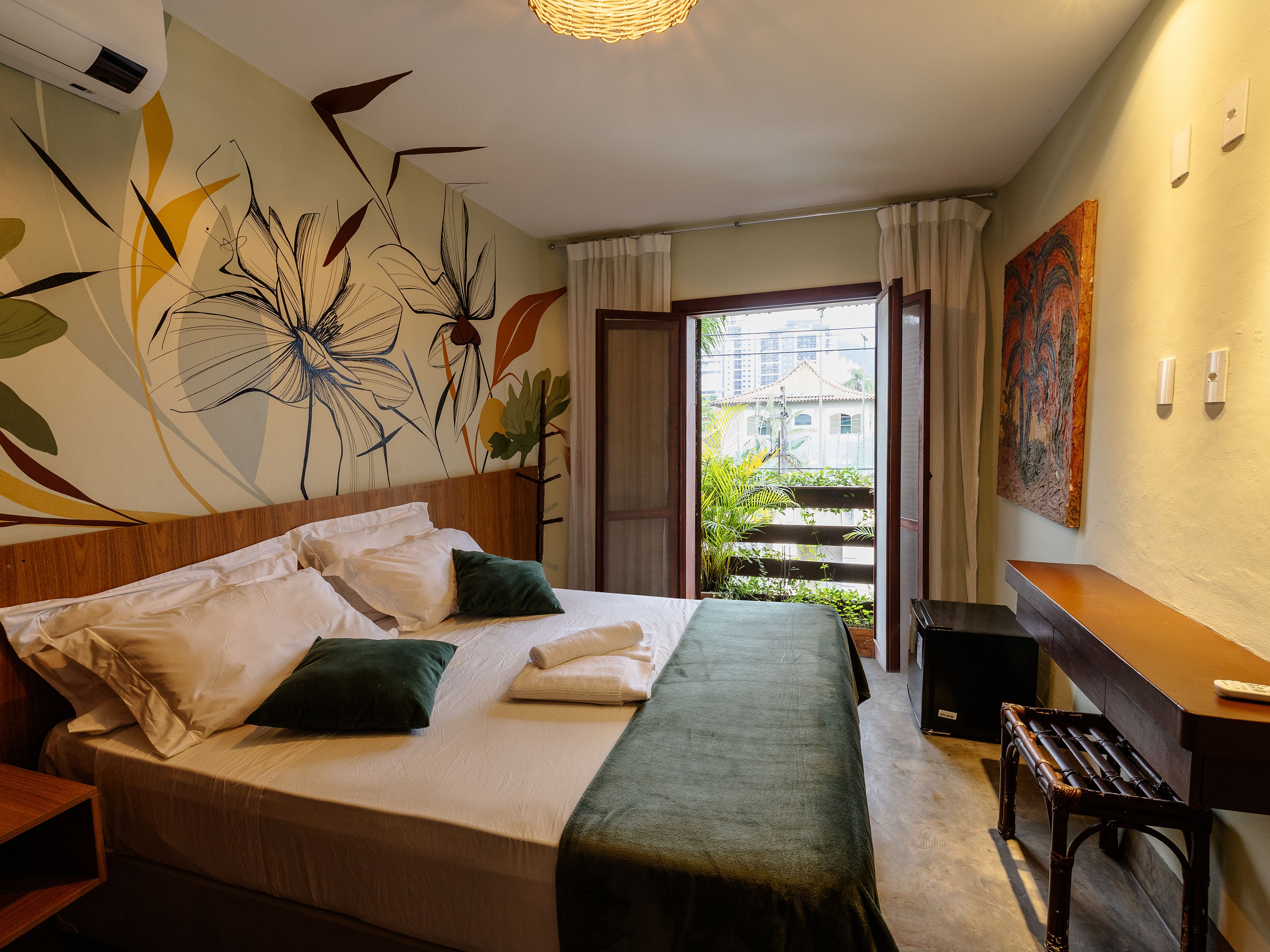A hotel room with a bed, a desk, a chair, a painting, and a view of buildings through the window.