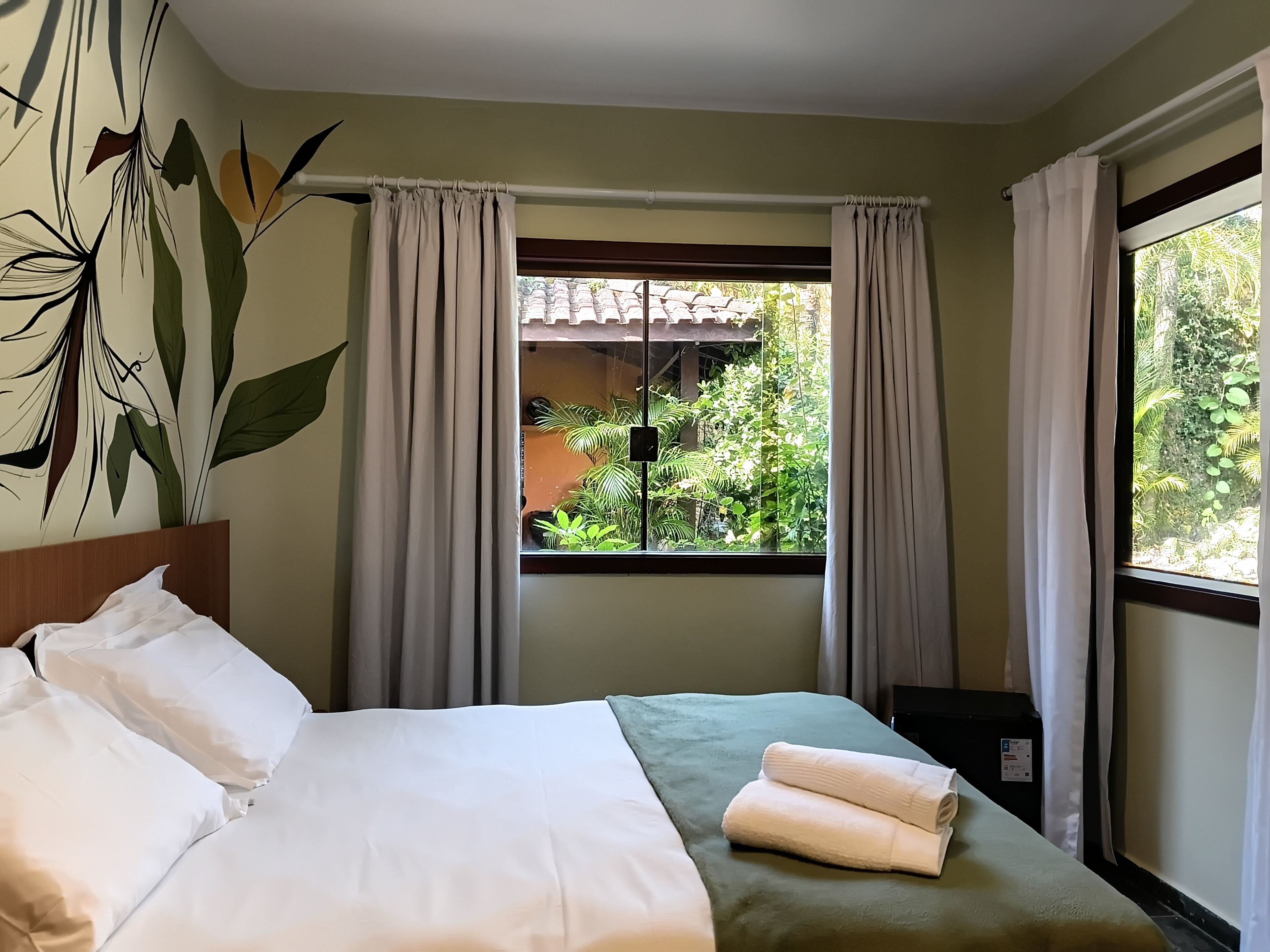 A hotel room with a bed, a window with curtains, a view of greenery outside, and a wall with a plant mural.