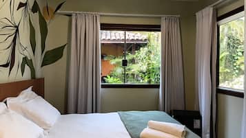 A hotel room with a bed, a window with curtains, a view of greenery outside, and a wall with a plant mural.