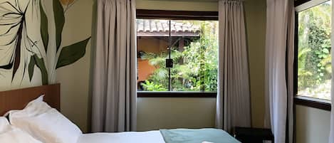 A hotel room with a bed, a window with curtains, a view of greenery outside, and a wall with a plant mural.