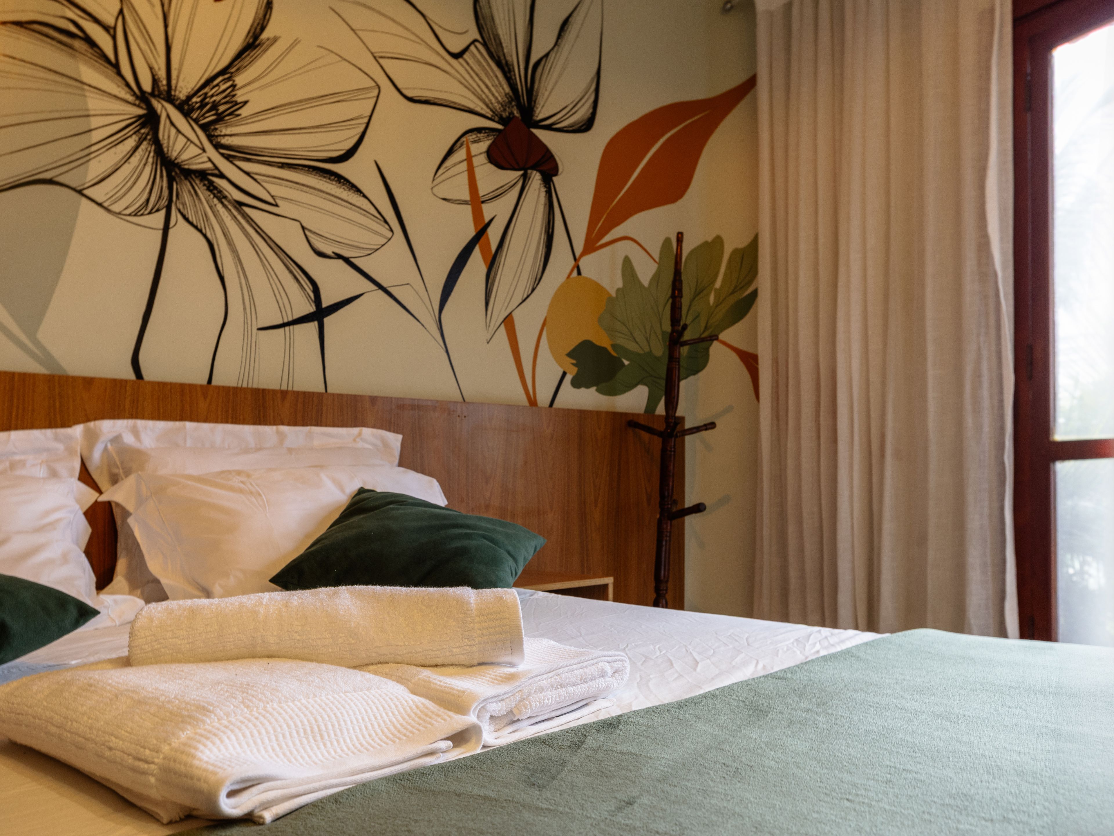 Cama bem arrumada com lençóis brancos e verdes, cabeceira de madeira e parede com um mural floral.