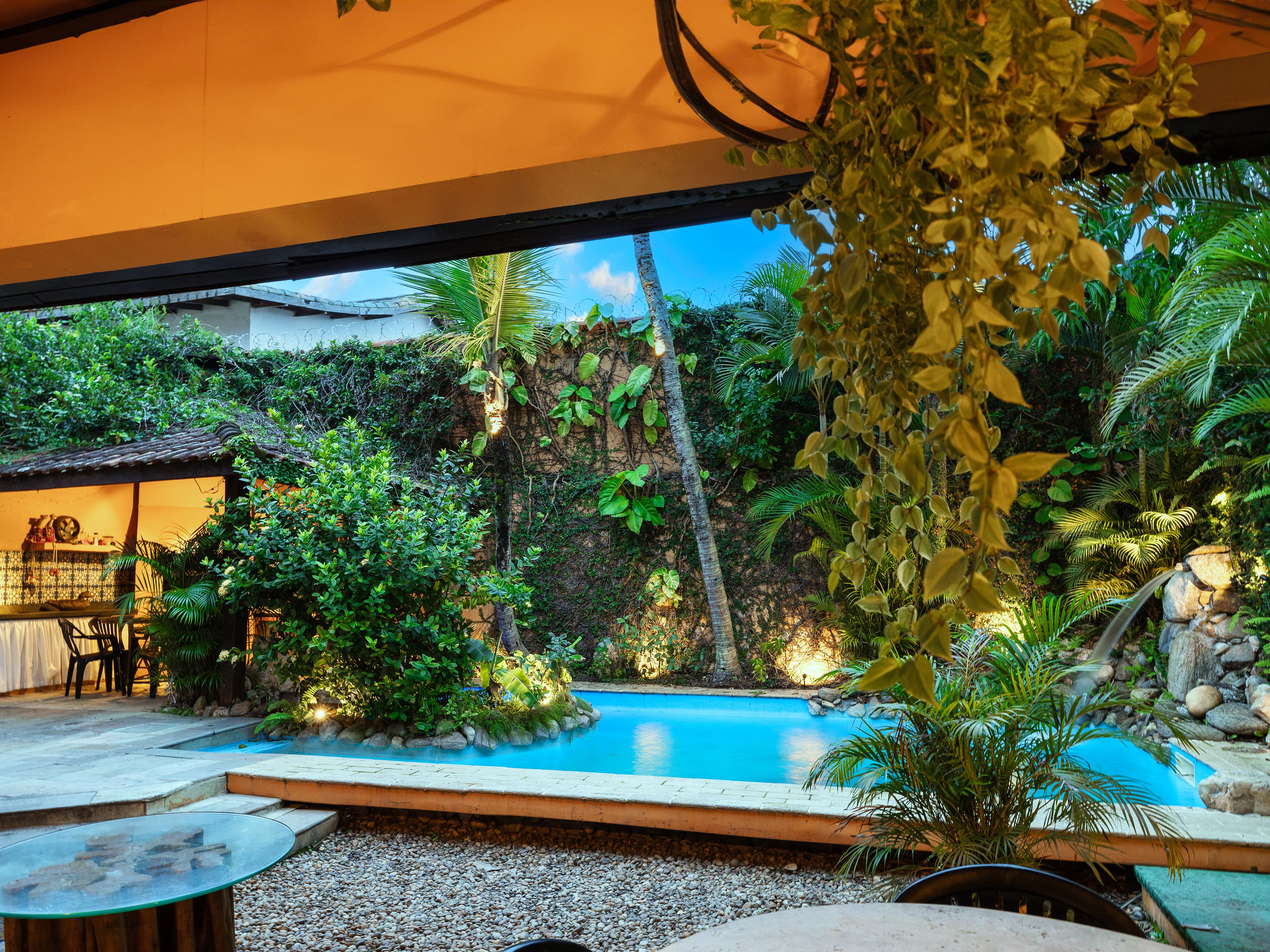 Un área de piscina con muro de piedra, plantas tropicales y una mesa con sillas.