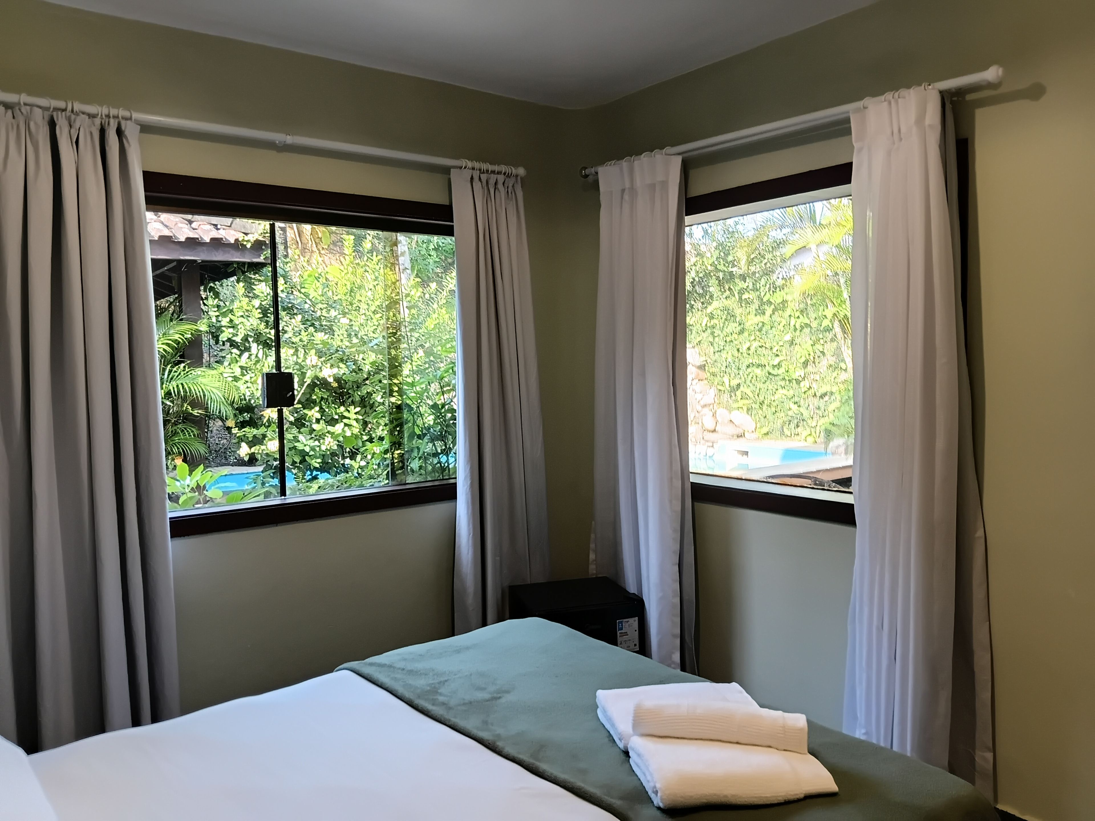 Habitación de hotel con cama, dos ventanas con cortinas, vista a zonas verdes y una piscina.