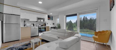 A modern living room with a white sofa, a small coffee table, and a view of a forest through large windows.