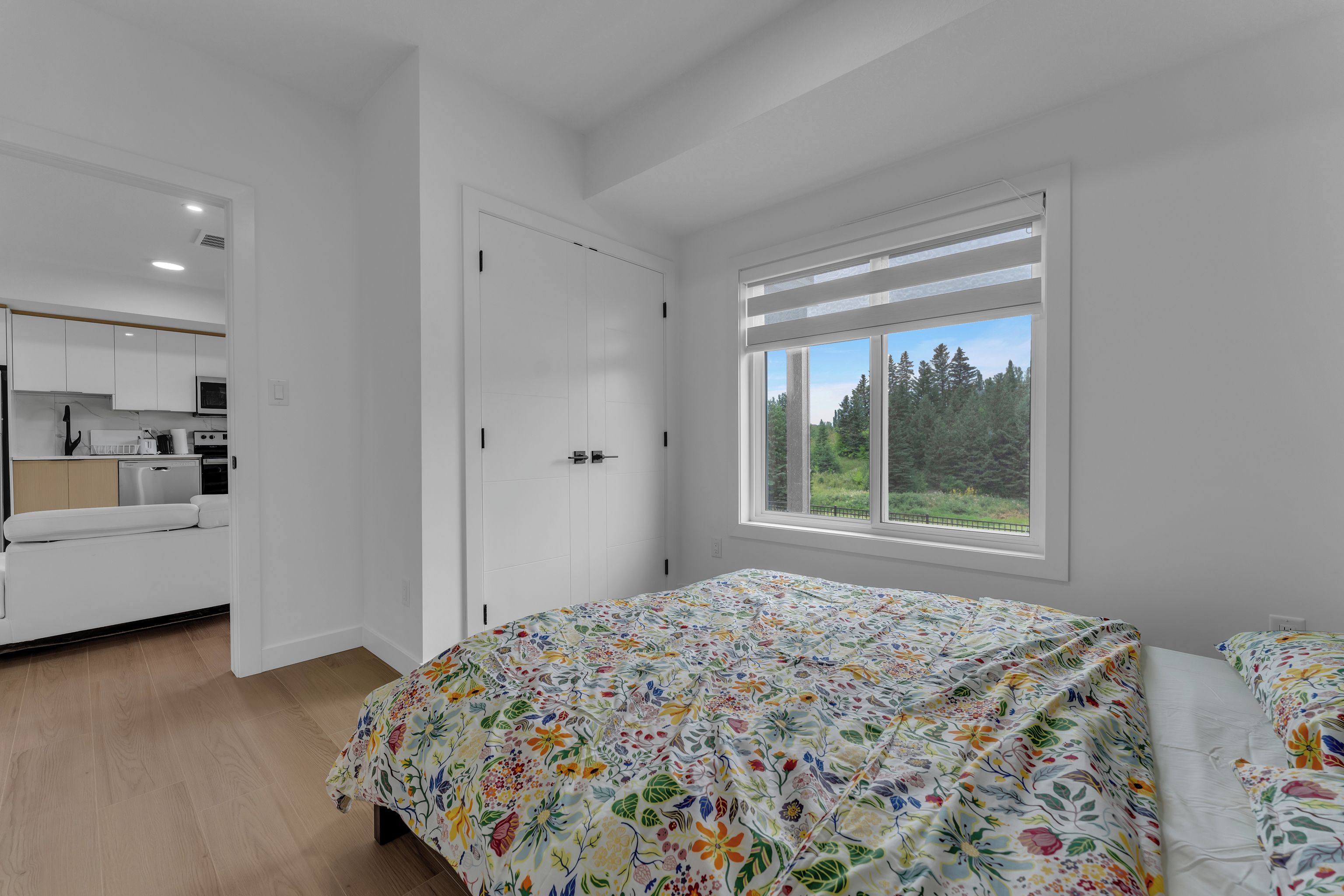 A bedroom with a bed, a window with a view of trees, and a kitchenette in the adjacent room.