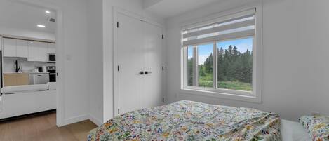 A bedroom with a bed, a window with a view of trees, and a kitchenette in the adjacent room.