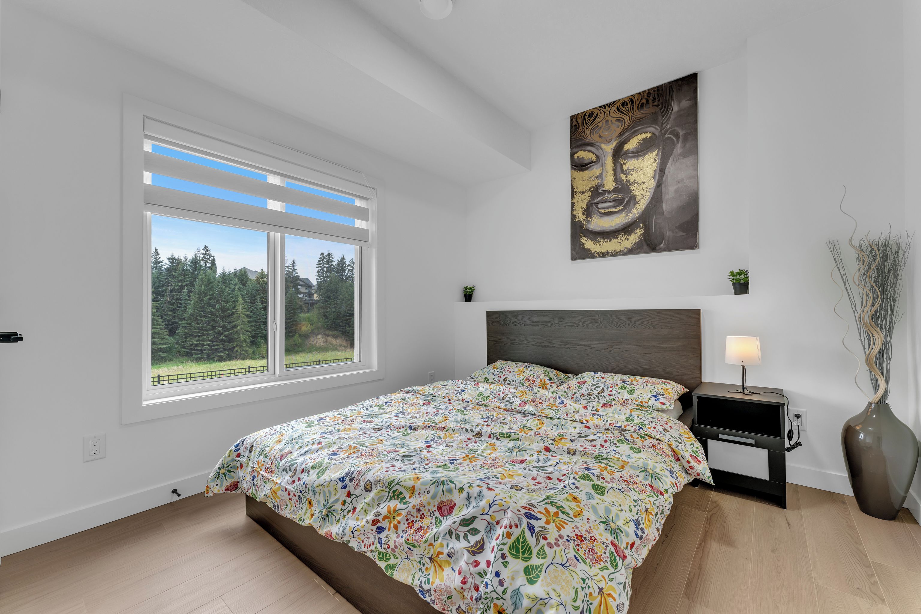 A bedroom with a large window, a bed with a floral bedspread, a nightstand with a lamp, and a wall-mounted artwork.