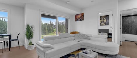 En moderne stue med en hvid hjørnesofa, et spisebord med stole, en potteplante og et stort vindue med udsigt til træer og bygninger.