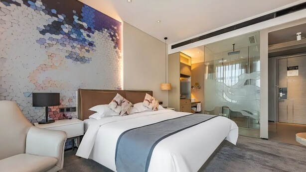 A modern hotel room with a large bed, a seating area, a glass partition, and a wall art piece.