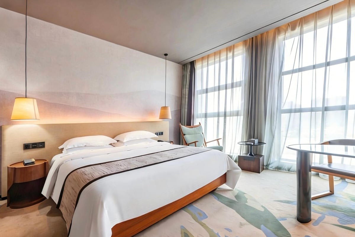 Habitación de hotel moderna con una cama grande, lámparas de noche, una mesita y vistas al exterior a través de cortinas translúcidas.