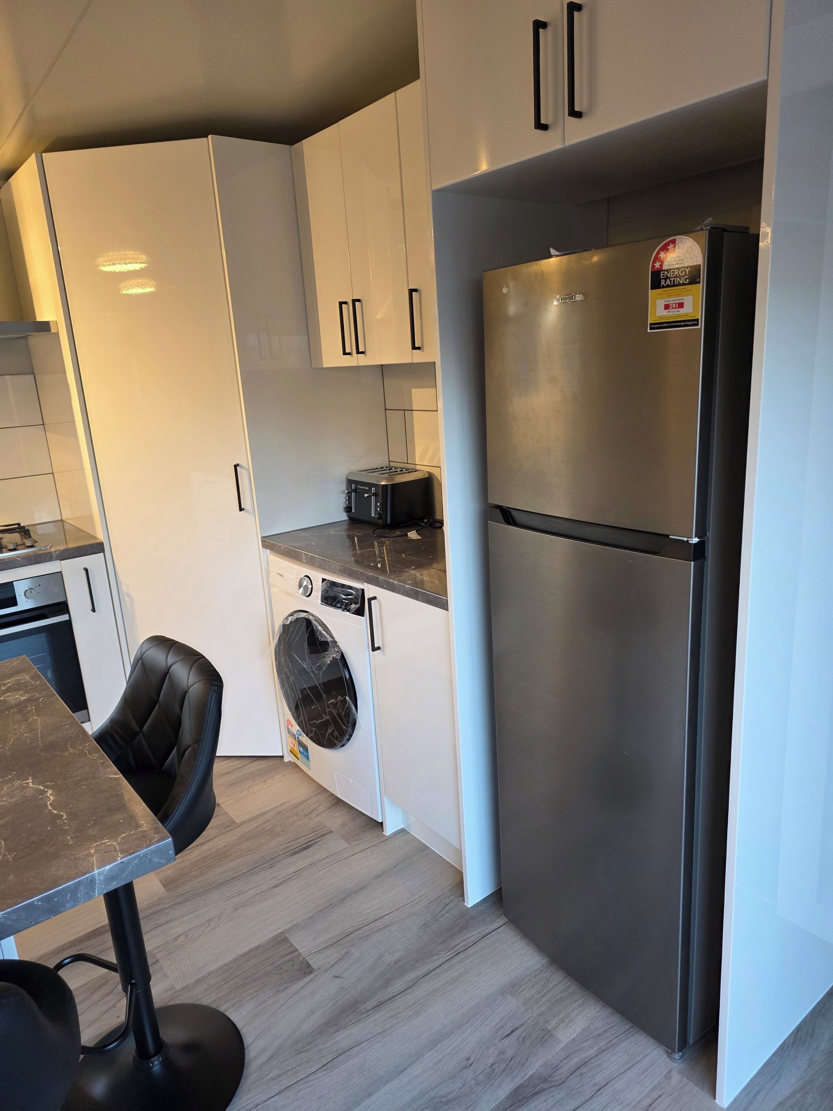 A compact kitchen with a built-in washing machine, a stainless steel refrigerator, and a black chair at a marble countertop.