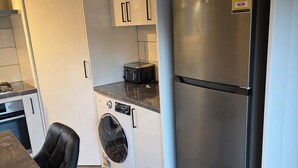 A compact kitchen with a built-in washing machine, a stainless steel refrigerator, and a black chair at a marble countertop.