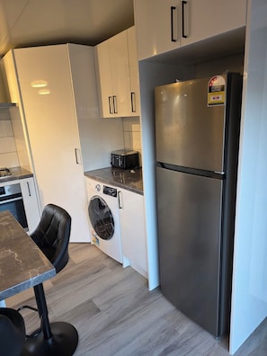 A compact kitchen with a built-in washing machine, a stainless steel refrigerator, and a black chair at a marble countertop.