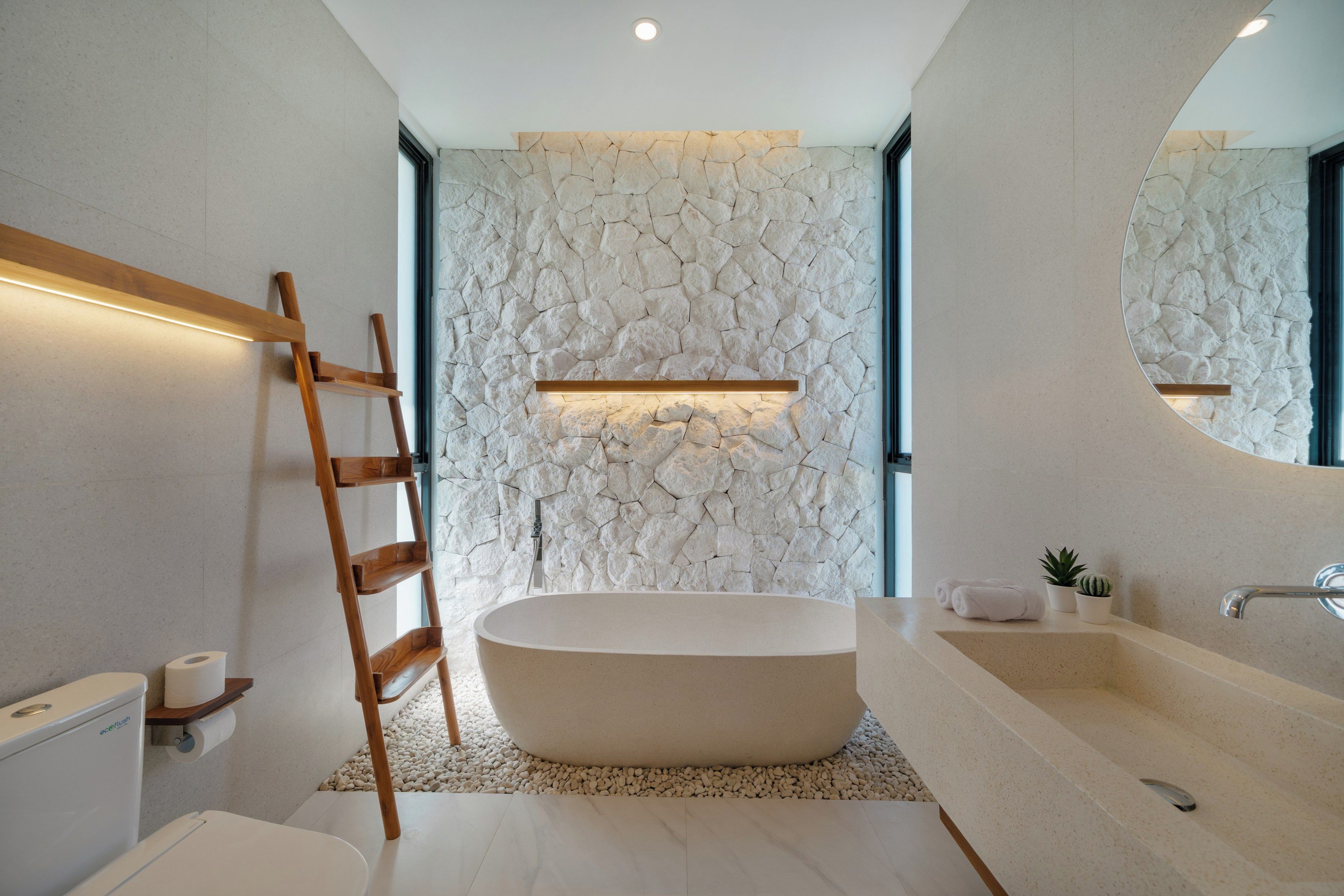 A modern bathroom with a freestanding bathtub, a wooden ladder, a stone wall, and a large mirror.