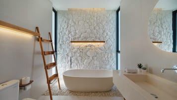 A modern bathroom with a freestanding bathtub, a wooden ladder, a stone wall, and a large mirror.