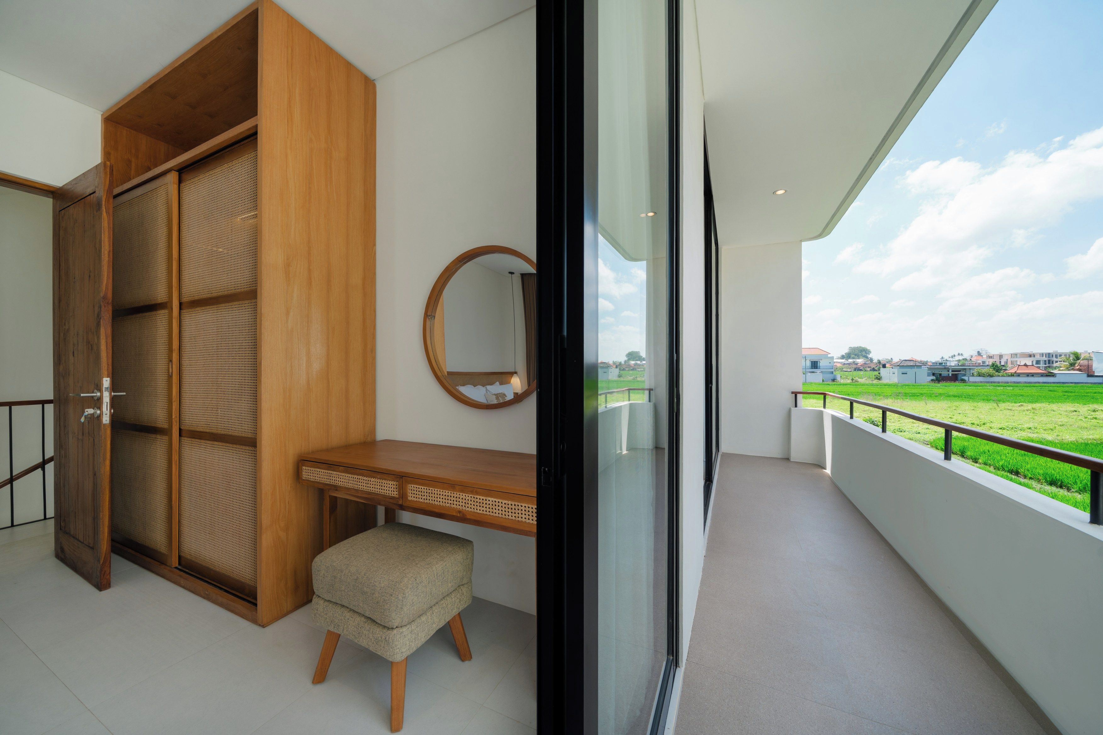 Ein Balkon mit Blick auf ein grünes Feld und Häuser, ein Holzschrank, ein runder Spiegel und ein kleiner Hocker.