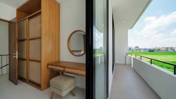 Ein Balkon mit Blick auf ein grünes Feld und Häuser, ein Holzschrank, ein runder Spiegel und ein kleiner Hocker.