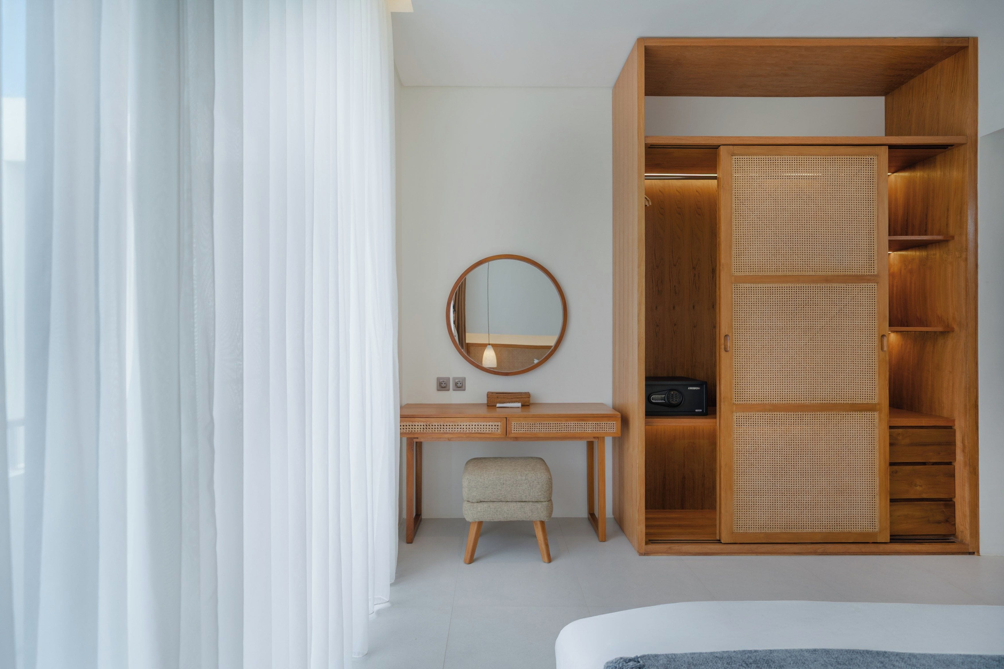 Une chambre d’hôtel avec une armoire en bois, un bureau avec un miroir, un petit tabouret et un lit.