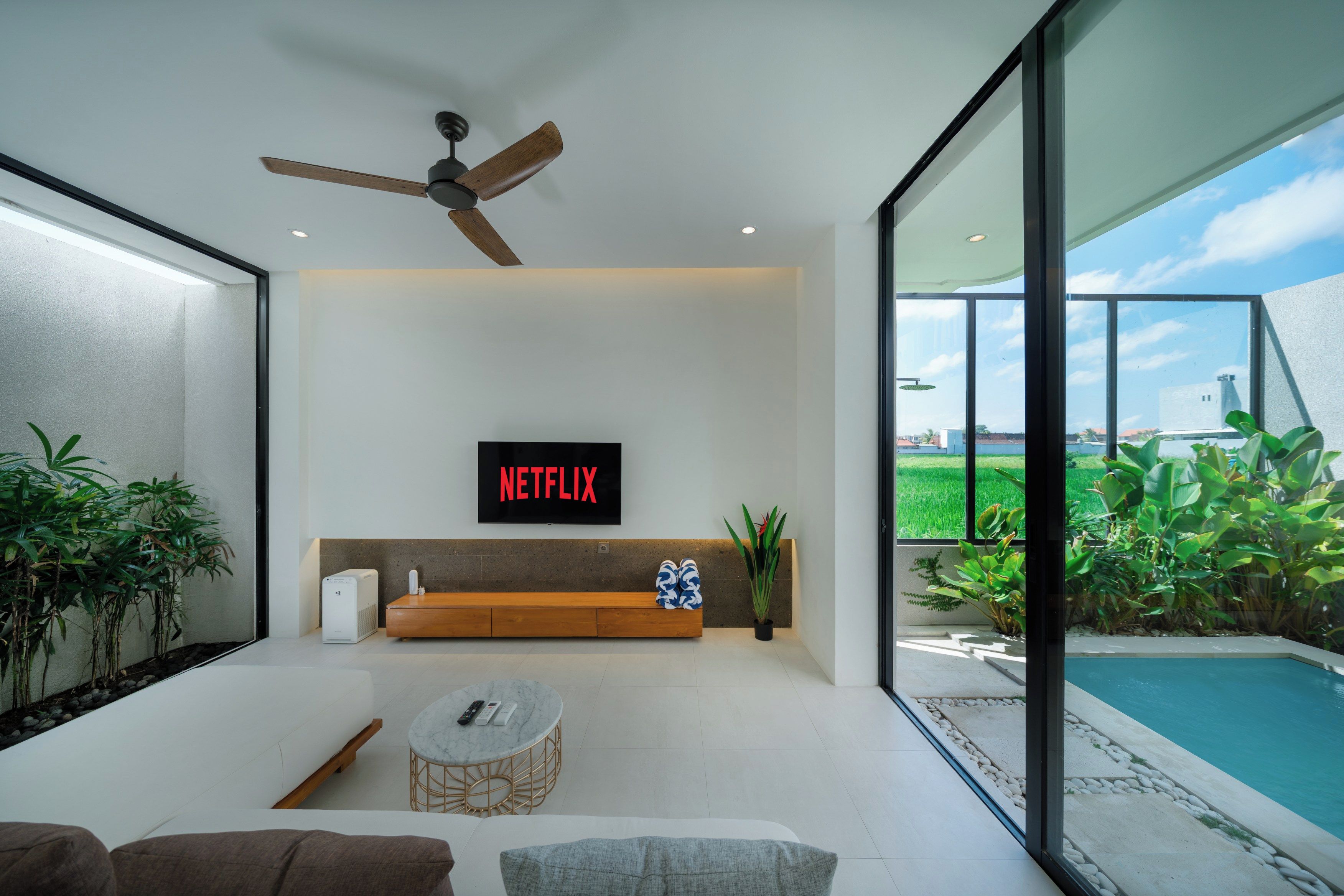 Un salon moderne avec un ventilateur de plafond, un logo Netflix, un canapé, une table basse et une vue sur une piscine.