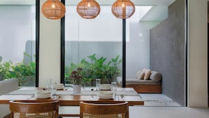 Uma sala de jantar com mesa posta para quatro pessoas, cercada por cadeiras de vime, e um espaço de estar com sofá e almofadas.