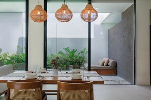 Un comedor con una mesa dispuesta para cuatro personas, rodeada de sillas de mimbre, y una zona de estar con un sofá y cojines.