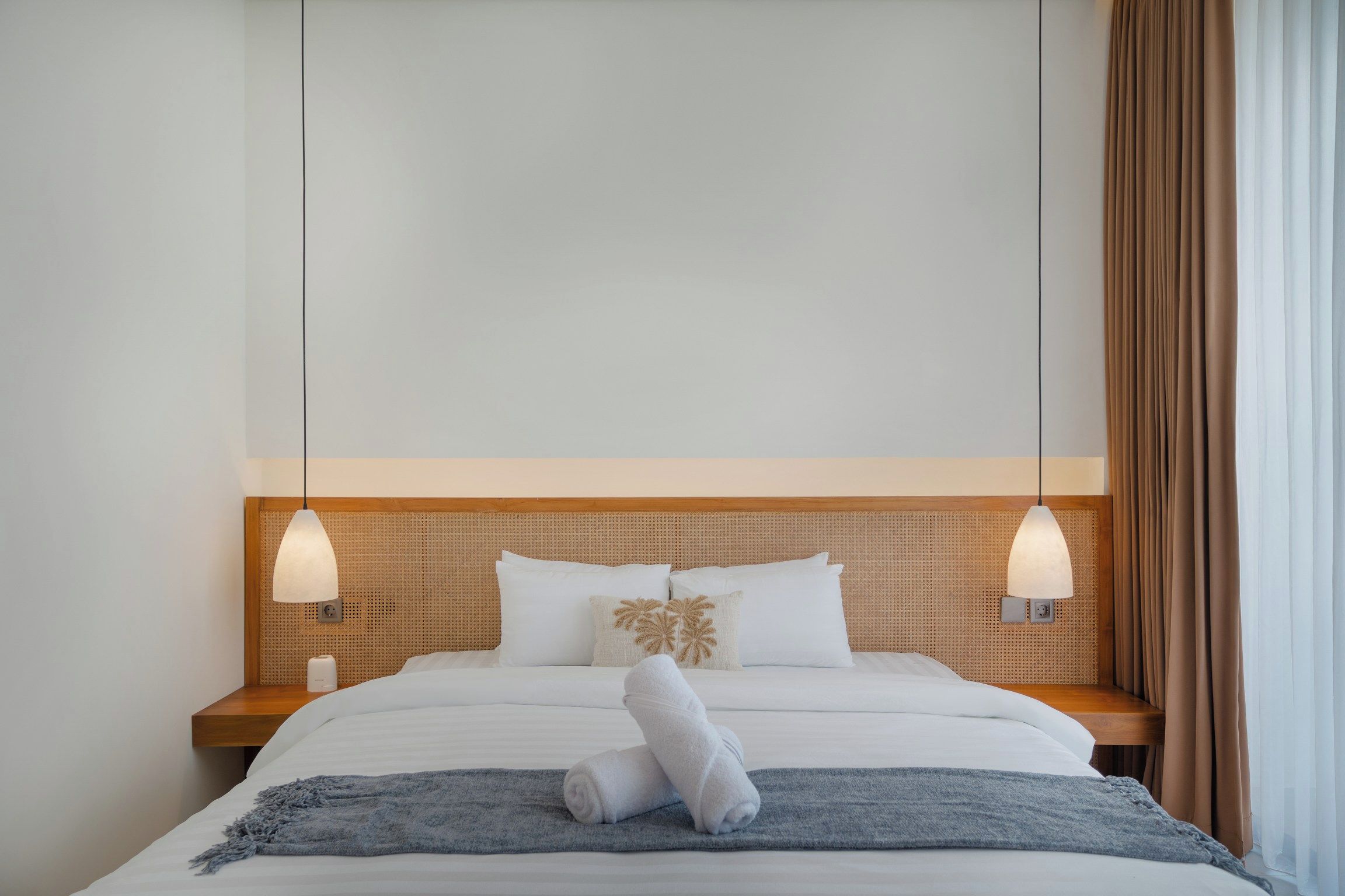 A hotel room with a bed, two bedside lamps, a wooden headboard, and a window with curtains.