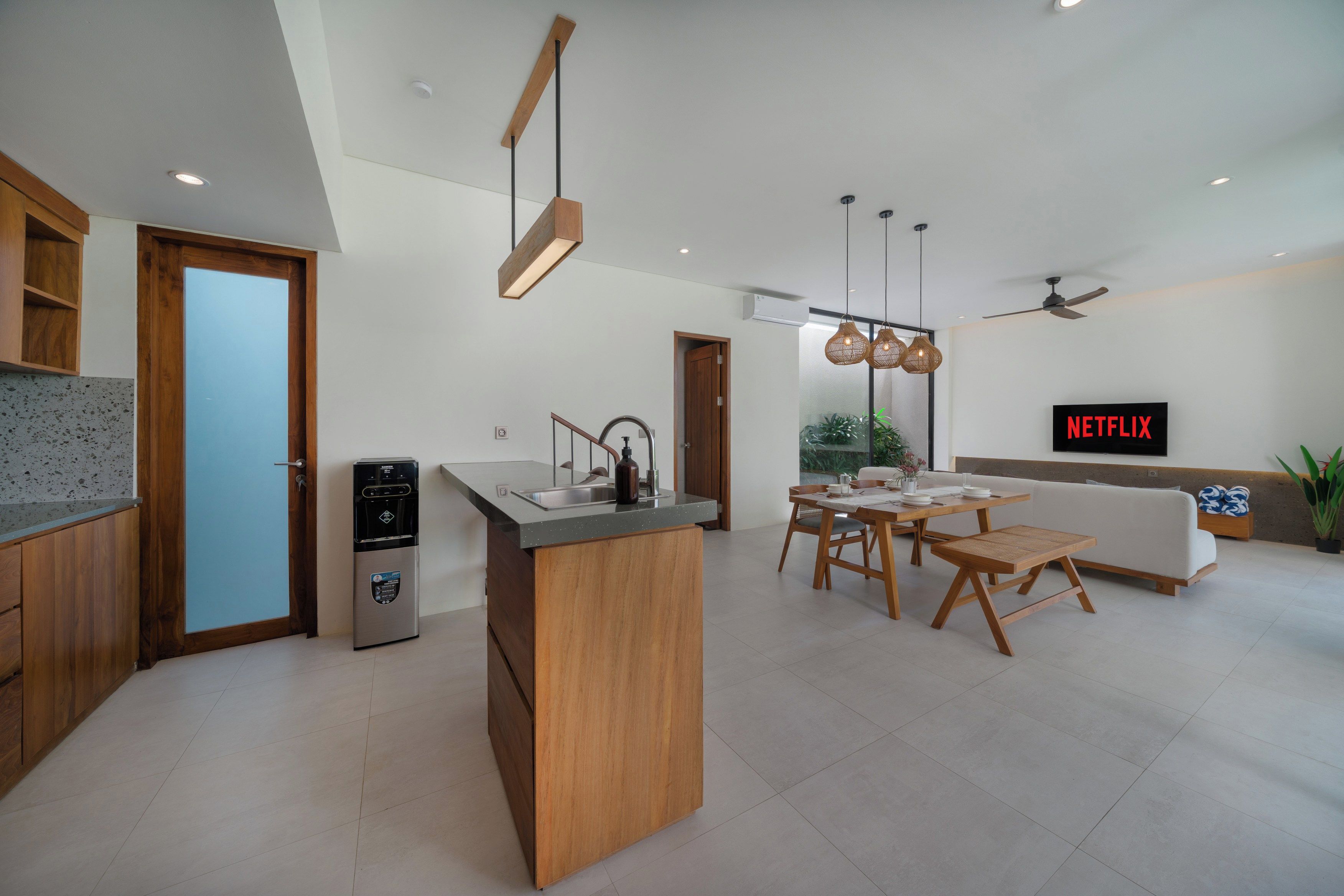 Una cocina moderna con una isla central, una zona de comedor con mesa y sillas de madera, un equipo de aire acondicionado empotrado y un televisor de pared mostrando el logotipo de Netflix.