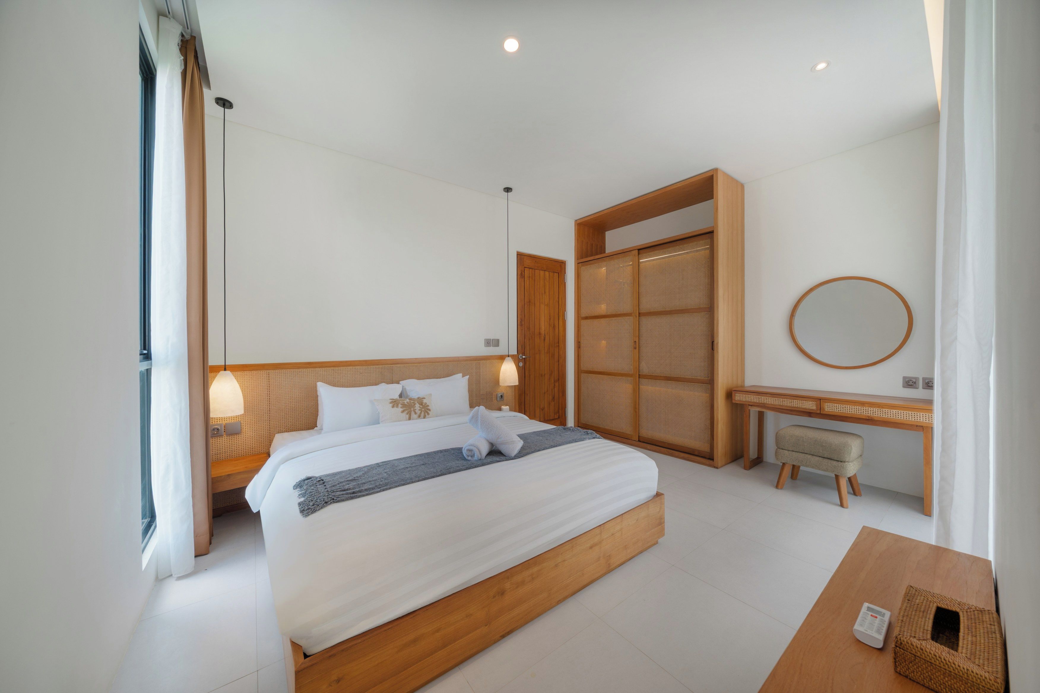 A modern bedroom with a large bed, bedside lamps, a wooden desk, a round mirror, and a woven wardrobe.