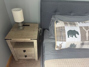 Ein Nachttisch aus Holz mit einer Lampe, ein Bett mit grauer Bettwäsche und ein gestepptes Kopfkissen mit Tiermustern.