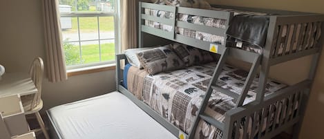 Un lit superposé avec un bureau et une chaise, une fenêtre avec des rideaux et une vue sur la verdure à l’extérieur.