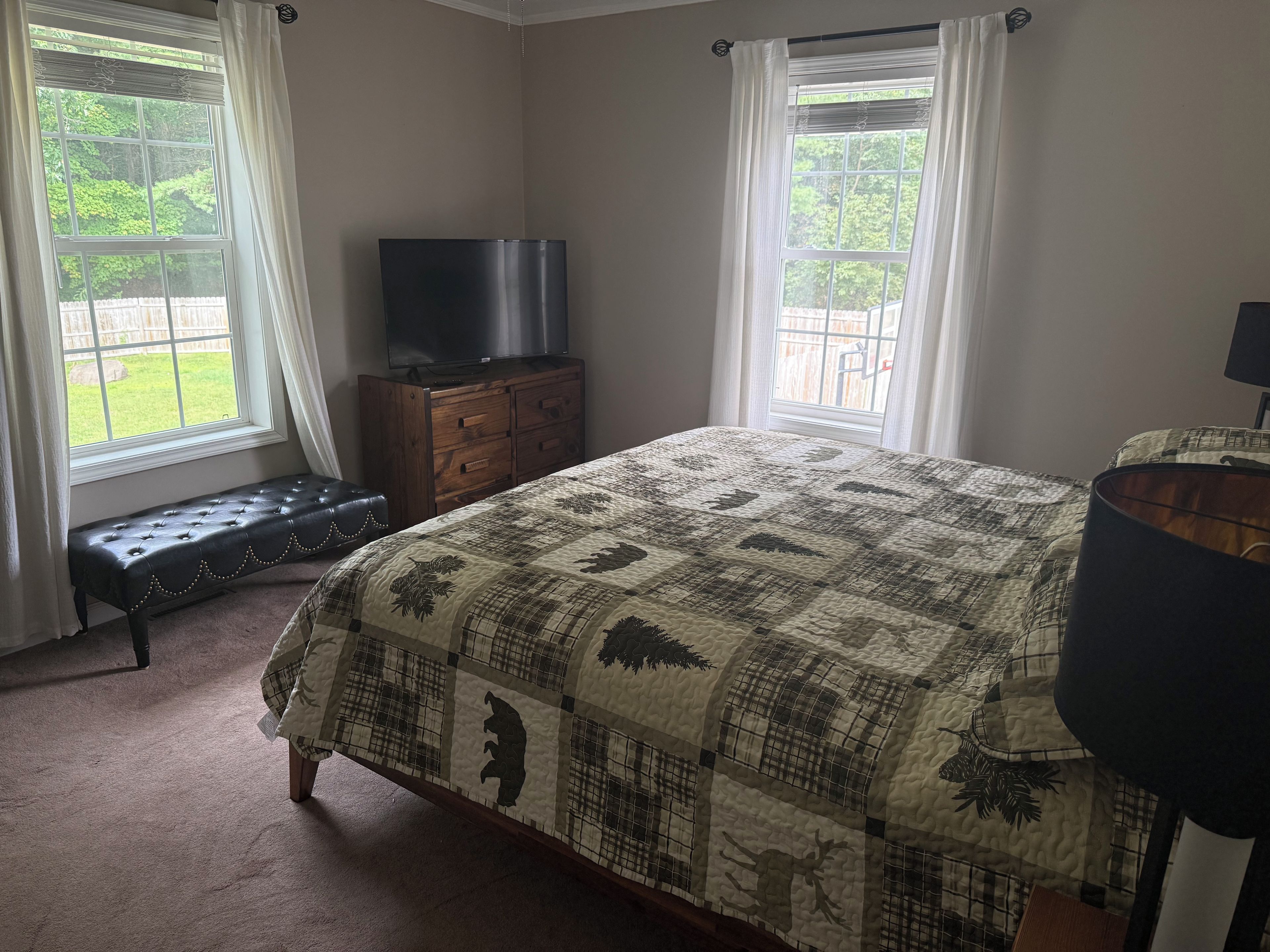 A bedroom with a large bed, a TV on a wooden dresser, and two windows with curtains.