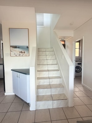 Eine Treppe mit weißer Geländer führt zu einer zweiten Etage, ein Küchenbereich mit Arbeitsplatte und Schränken sowie ein Hauswirtschaftsraum mit Waschmaschine.