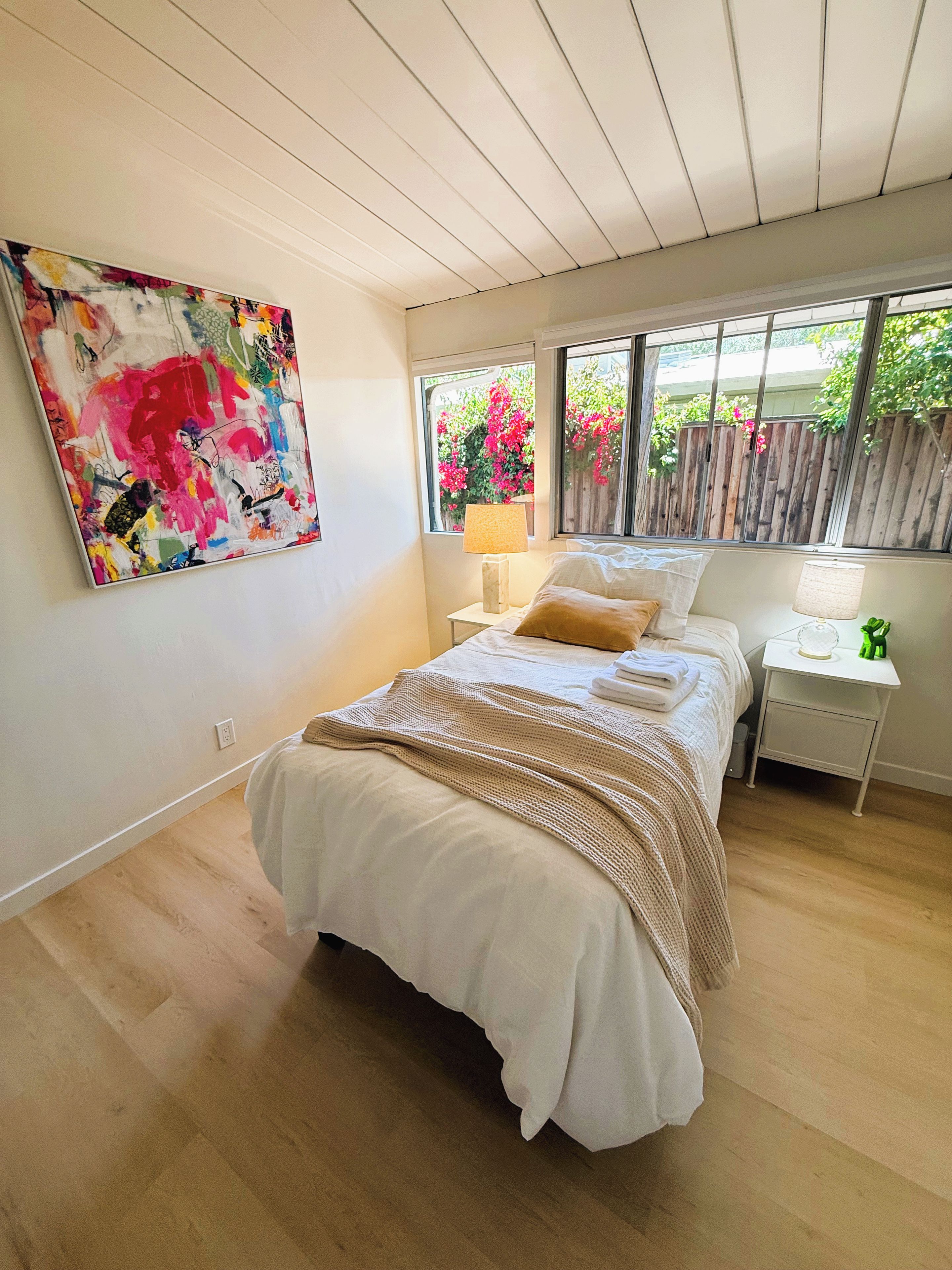 Een slaapkamer met een bed, een nachtkastje, een lamp, een raam met uitzicht op bloemen en een groot abstract schilderij aan de muur.