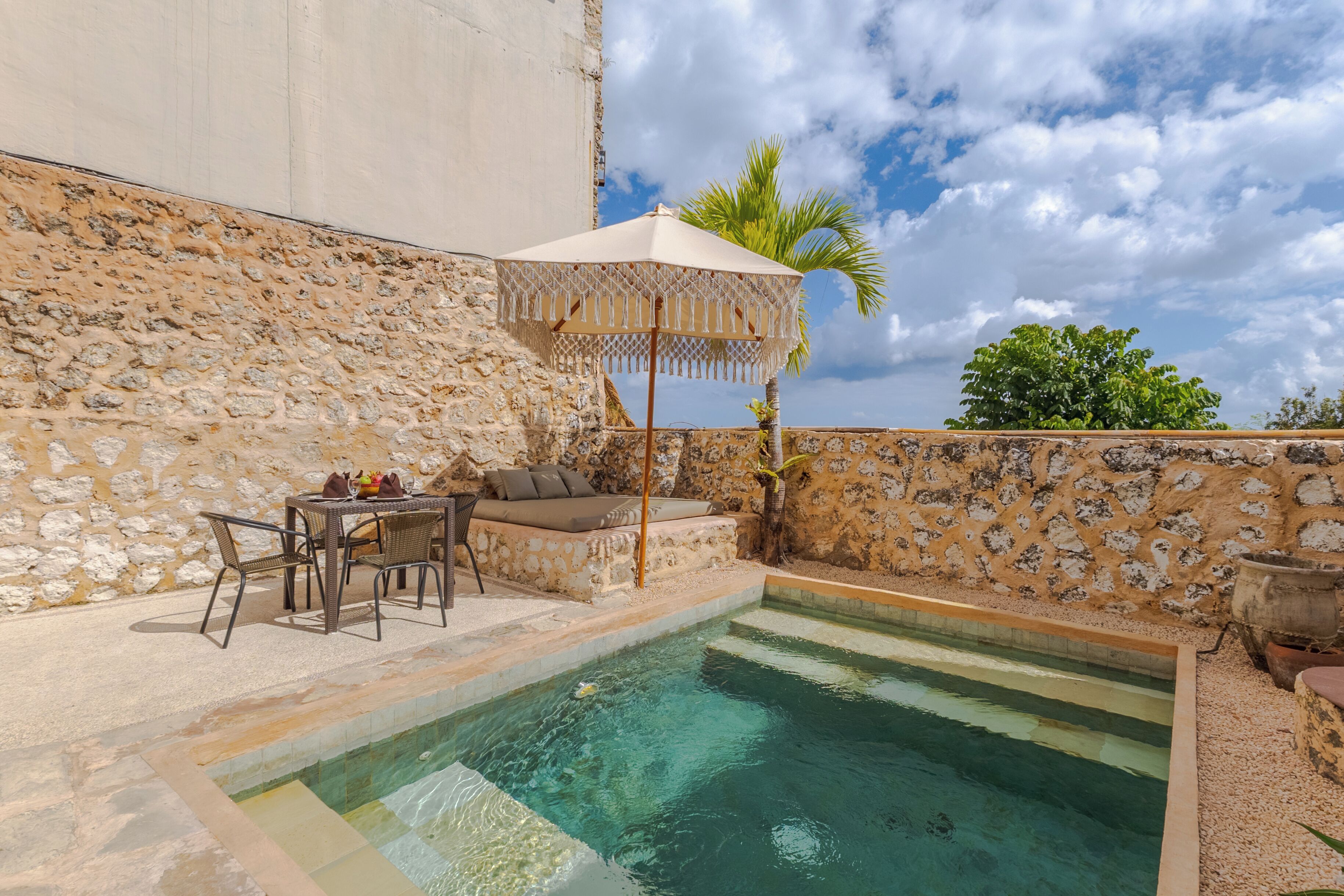 A stone-walled outdoor area with a pool, a table with chairs, and a palm tree.