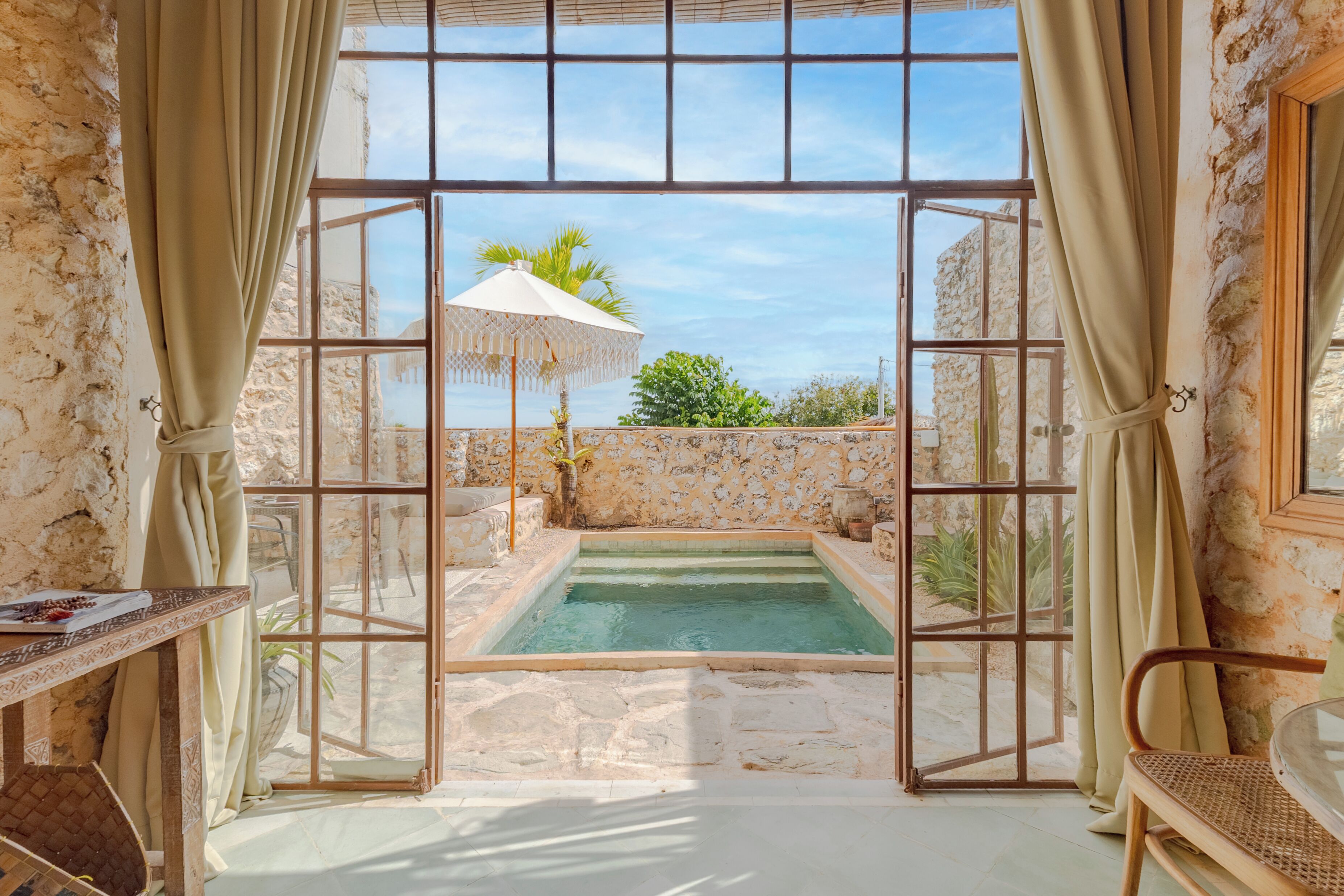 Una habitación con pared de piedra, una ventana con cortinas, una piscina pequeña y una mesa con una silla.