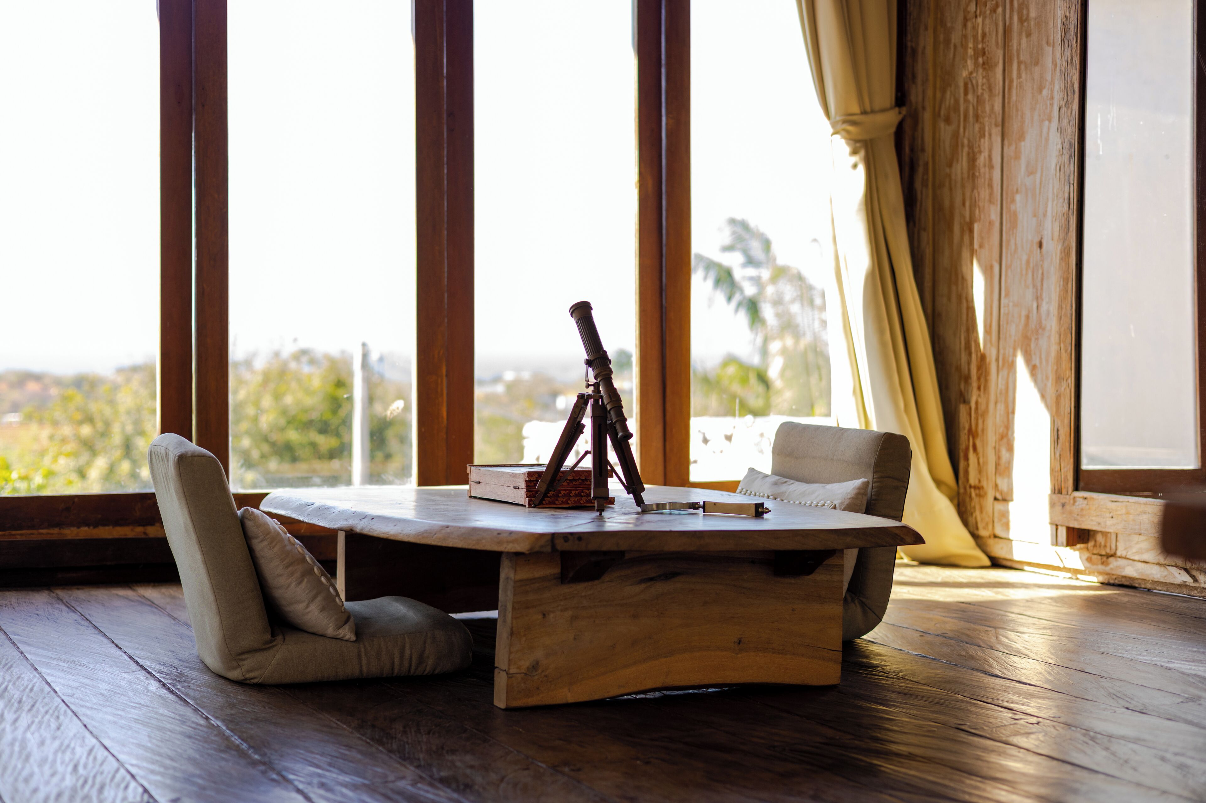 Een houten tafel met een statief en een doos, twee stoelen, en uitzicht door grote ramen.