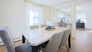 A modern dining room with a marble table, grey upholstered chairs, and a kitchen in the background. - Renovated Lakefront 5BR • Near Colleges • Sunsets • Sleeps 12 (Worcester)