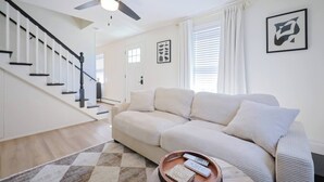 A modern living room with a staircase, a sofa, a coffee table with remotes, and a ceiling fan. - Renovated Lakefront 5BR • Near Colleges • Sunsets • Sleeps 12 (Worcester)