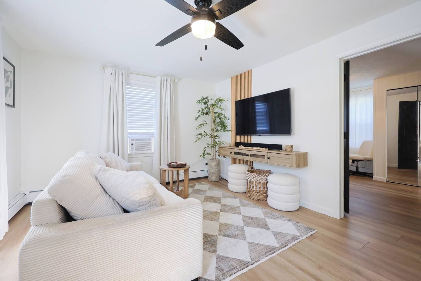 A modern living room with a sofa, a television, a ceiling fan, and a large window with curtains.