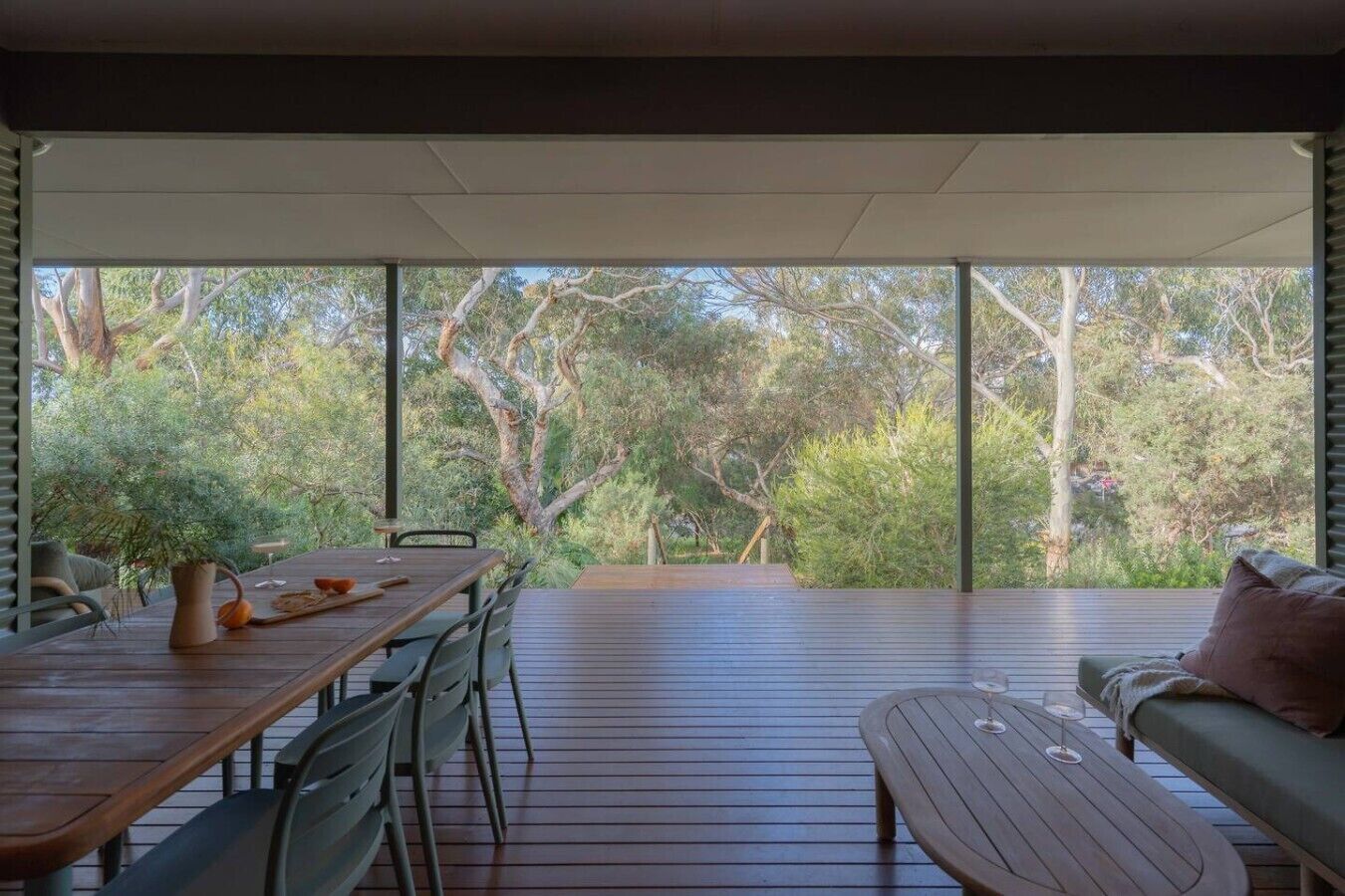 Un espace de restauration en plein air avec une table et des chaises, un banc et une vue sur les arbres à travers de grandes fenêtres.