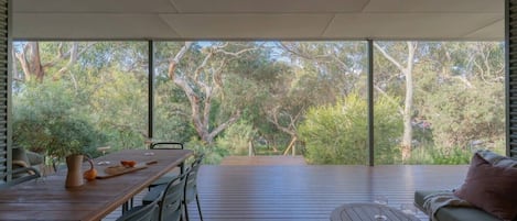Un espace de restauration en plein air avec une table et des chaises, un banc et une vue sur les arbres à travers de grandes fenêtres.
