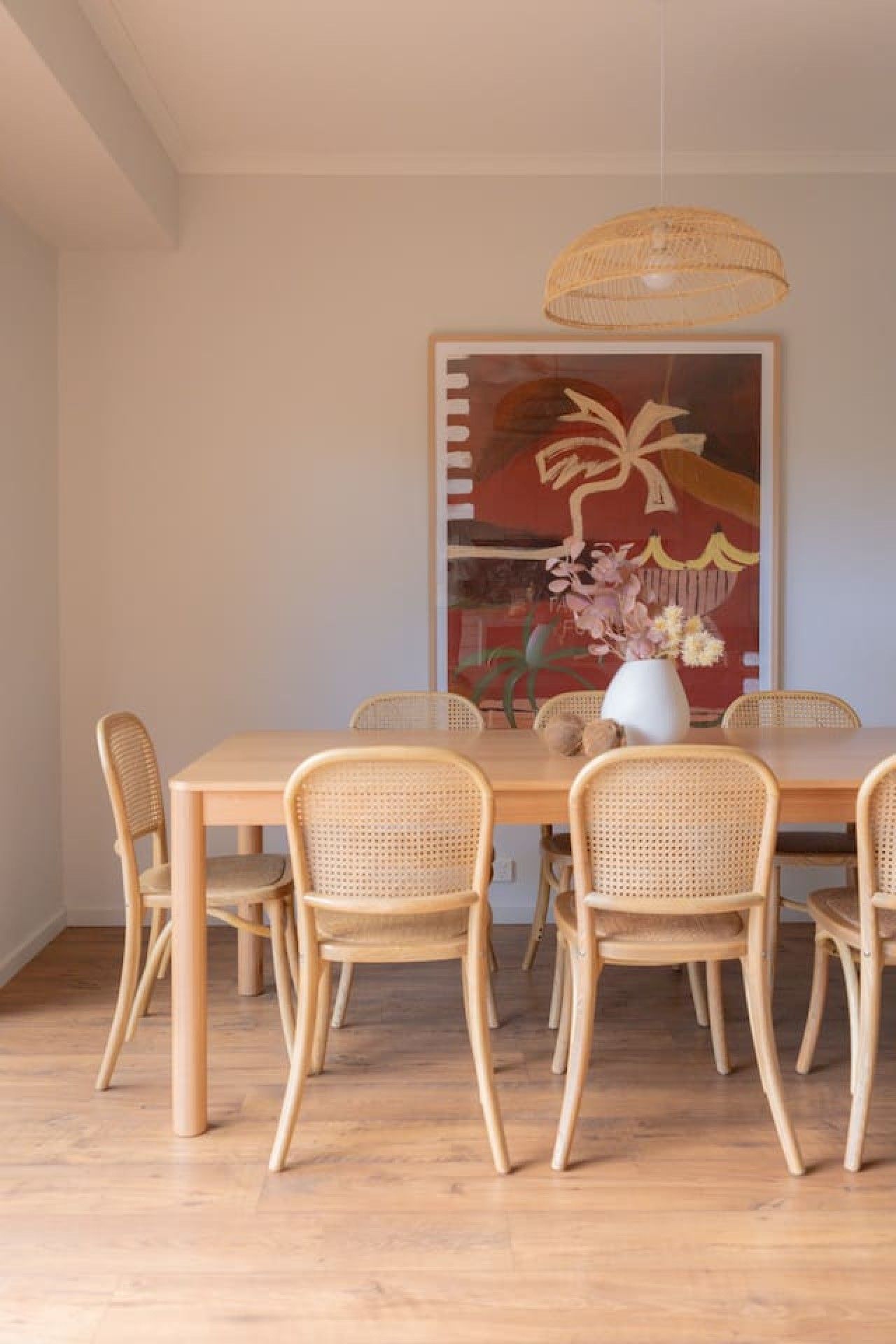 Een eetruimte met een houten tafel en rieten stoelen, een groot abstract schilderij aan de muur en een hanglamp.