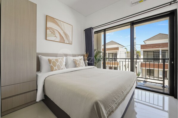 A modern bedroom with a large bed, a painting on the wall, and a view of neighboring buildings through a balcony.