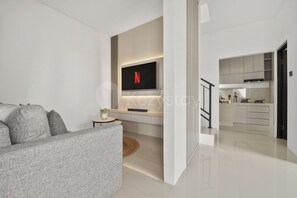 A modern living room with a grey sofa, a flat-screen TV mounted on the wall, and a kitchen area with white cabinets and a black countertop.