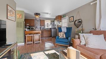 A living room with a sofa, a blue armchair, a television, and a kitchen in the background.