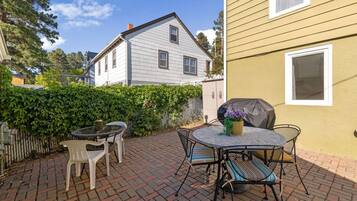 Une terrasse avec une table ronde et des chaises, un barbecue et une haie.