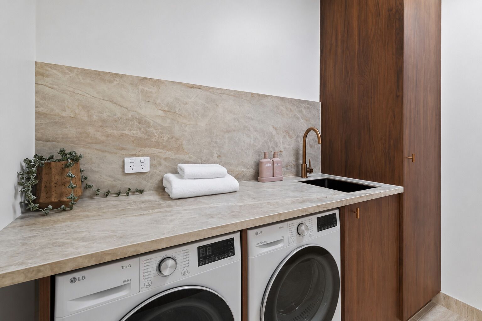 A laundry area with a washing machine, dryer, and a sink with a faucet. There are towels stacked on the counter and a potted plant.