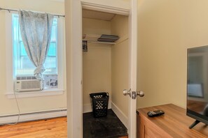 Um quarto com piso de madeira, uma TV sobre um móvel de madeira, uma porta com puxador redondo e uma janela com persianas.