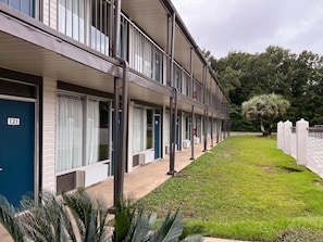 Fila de habitaciones de hotel con un paseo techado, un letrero con el número '121' y un pequeño césped con palmeras.